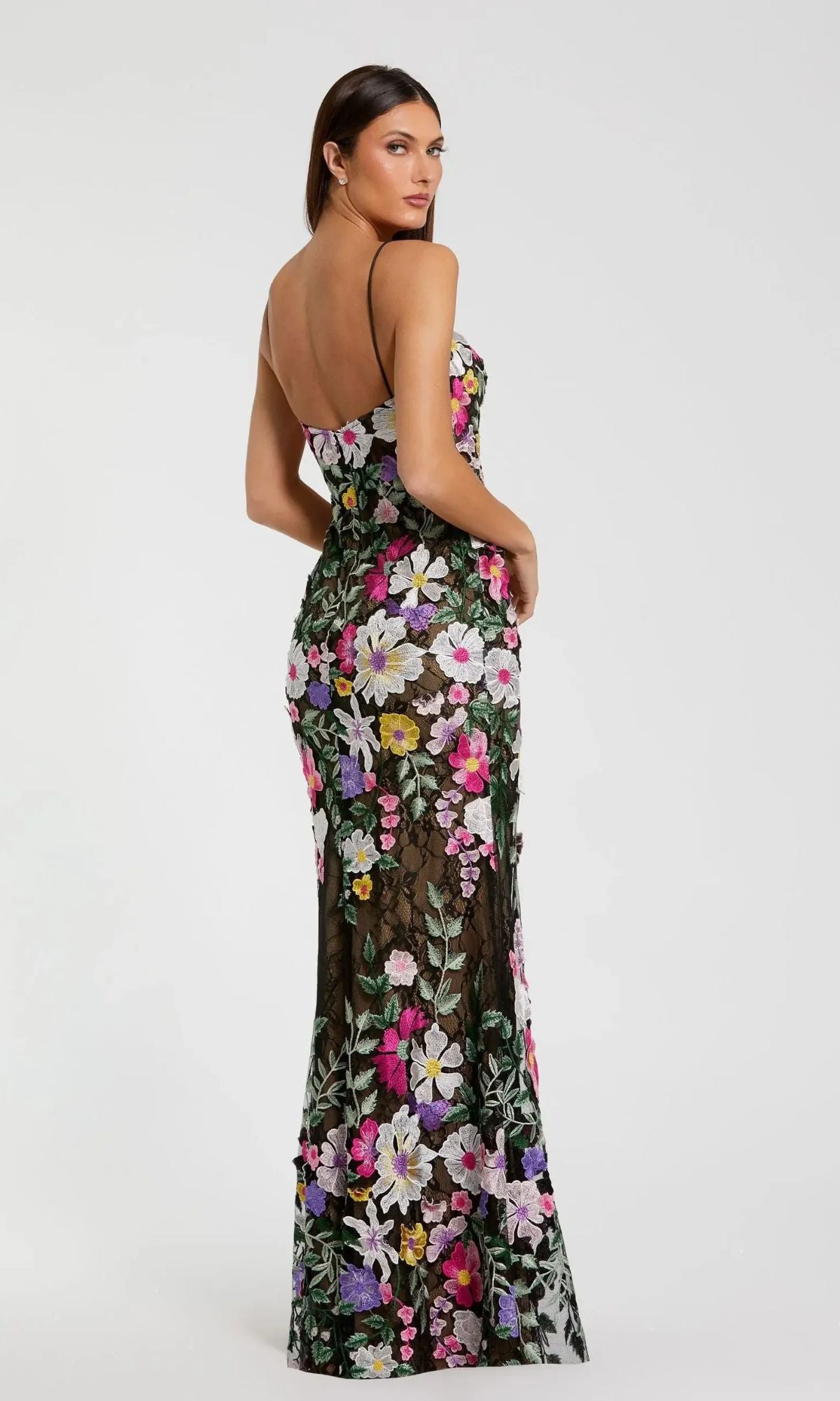 A woman faces away wearing the Mac Duggal 68897 long formal dress, a fitted black gown with thin straps and vibrant floral embroidery, its colorful patterns accenting the floor-length silhouette against a plain light background.